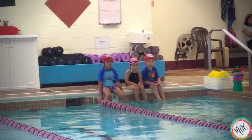 3 children in their red turtle 1 british swim school caps sitting on the side of the pool waiting for their turn at the lesson..