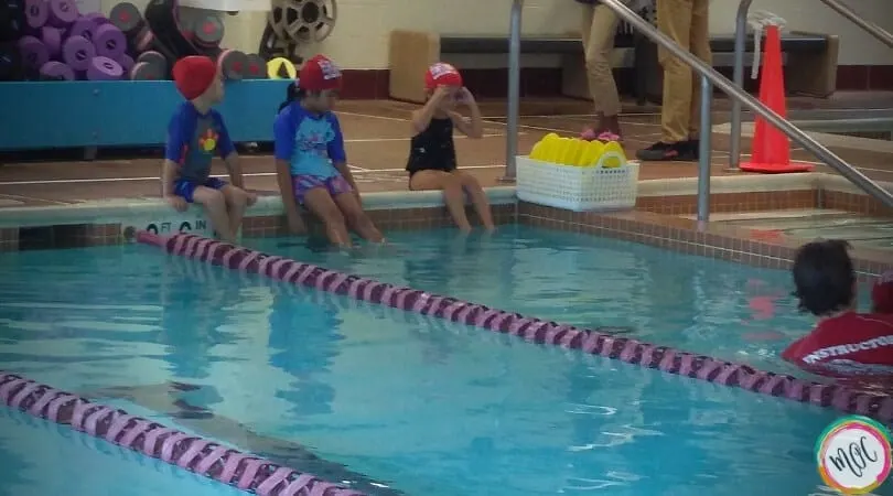 3 children sitting at the side of the pool in their red turtle 1 british swim school caps, waiting their turn for the lesson. One child getting her goggles on.