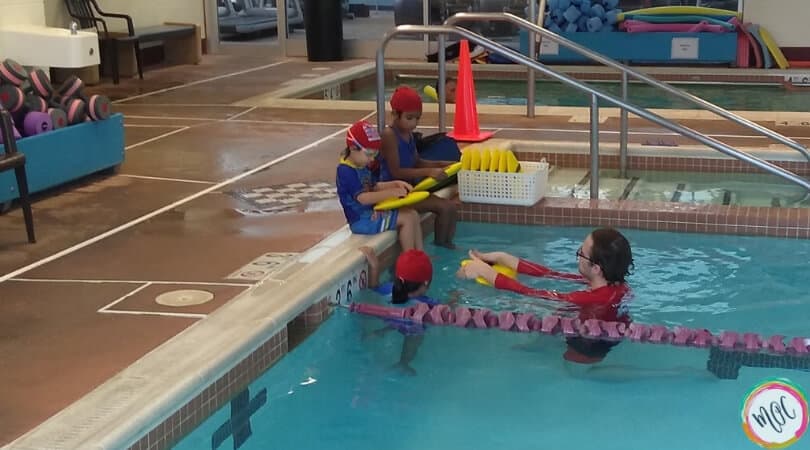 British Swim School- A Girl and Her Goggles turtle 1 level red swim caps for british swim school, children sitting at edge of the pool during class.